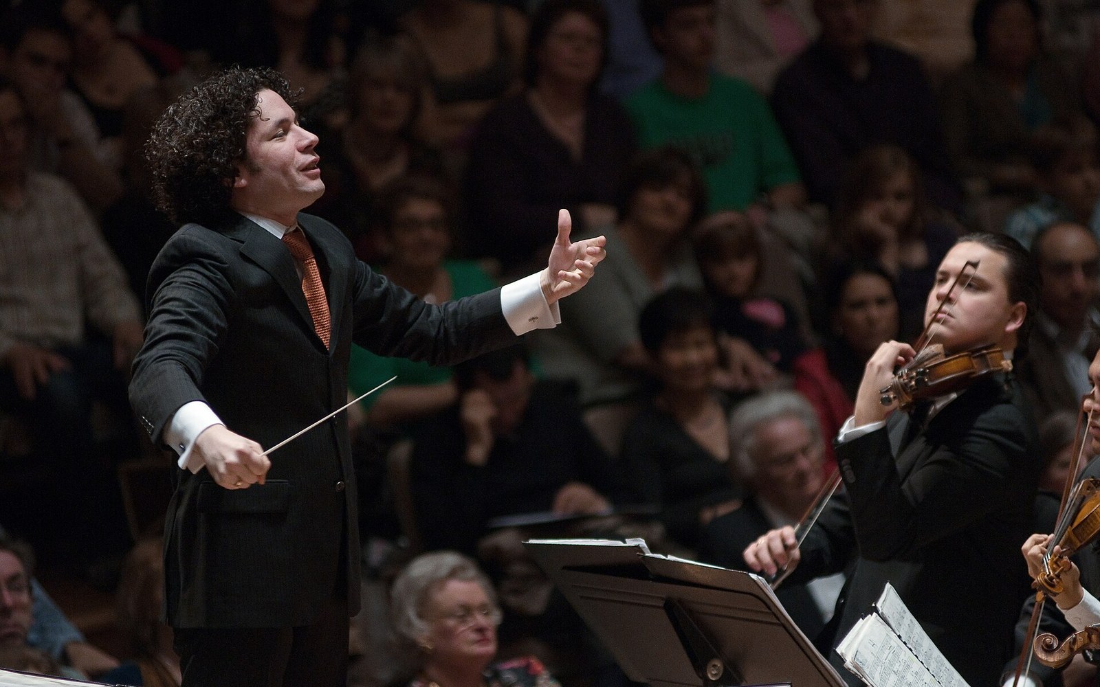 Gustavo Dudamel conducting the Simón Bolívar Symphony Orchestra, adult musicians visible in the mid-ground, mid-performance