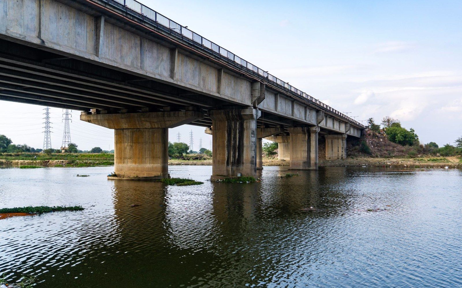 Concrete pillars support a long bridge crossing a calm river beneath a partly cloudy sky