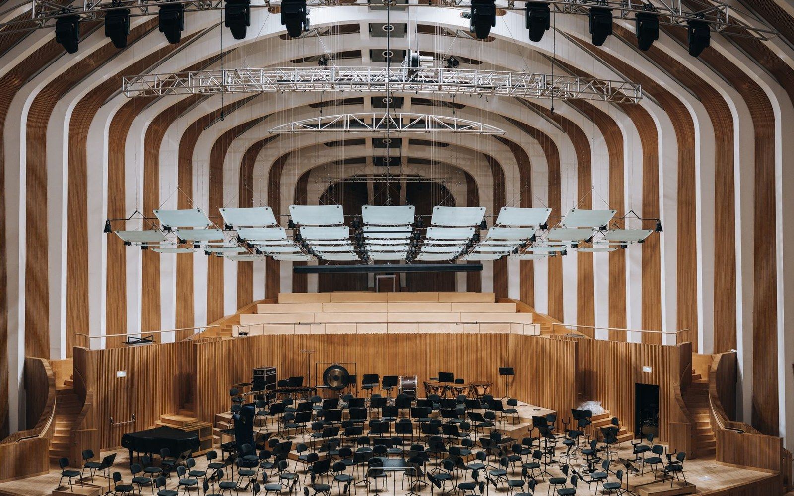 Concert hall stage with empty chairs beneath suspended acoustic panels and striped wooden ceiling