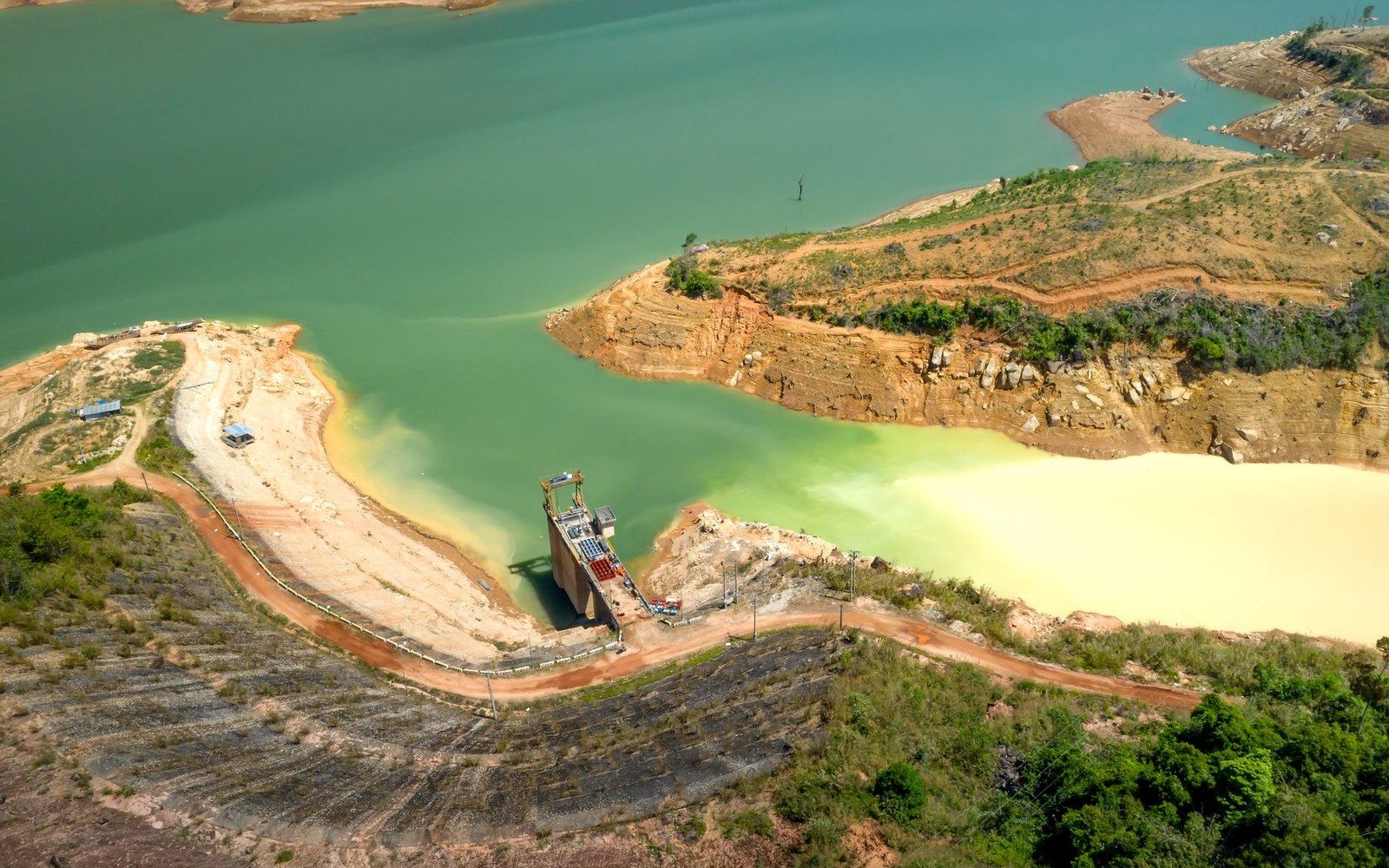 Aerial view of a dam structure between turquoise and murky yellow reservoir waters