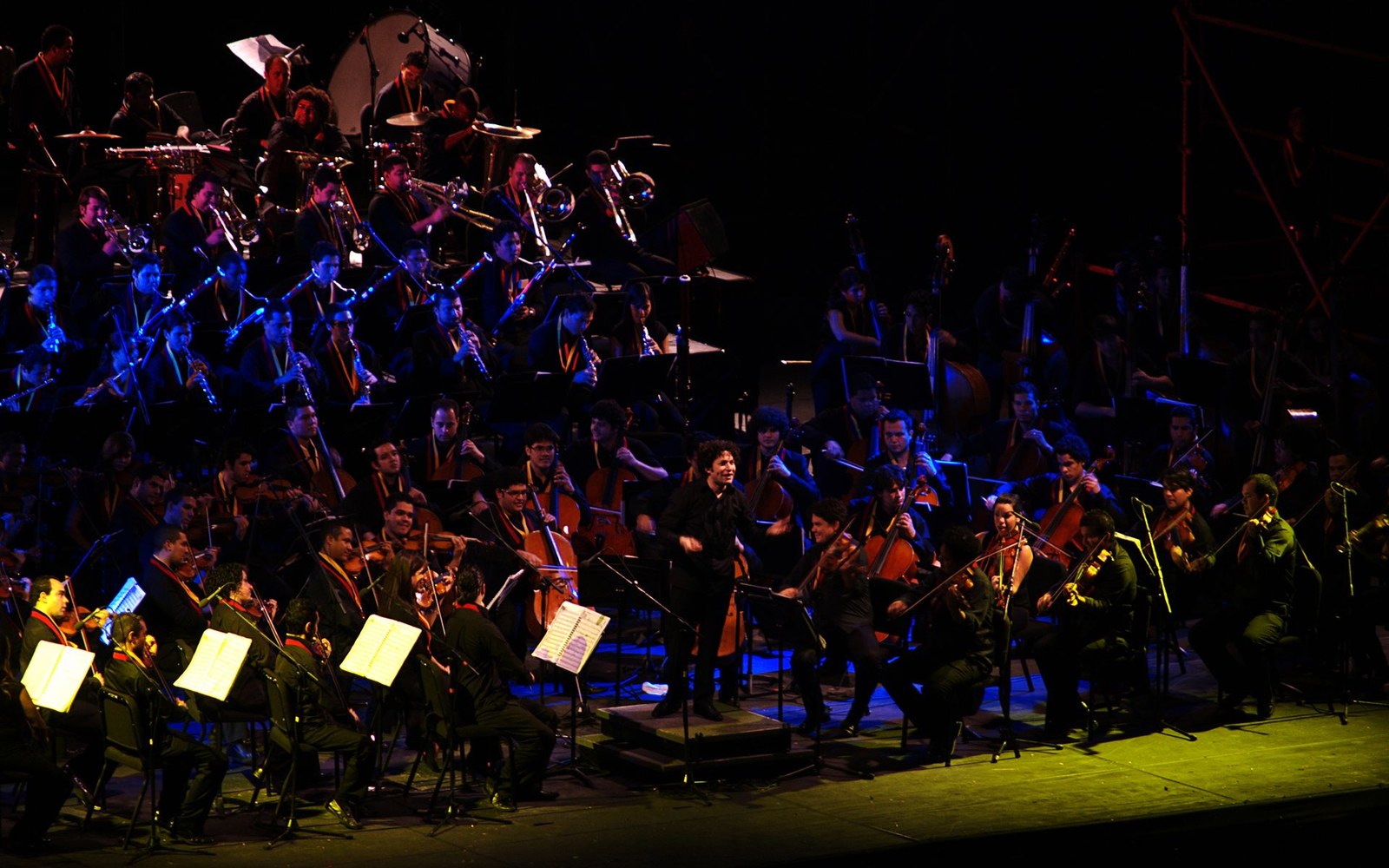 A conductor leads a large youth orchestra in a dramatic blue-lit performance on stage
