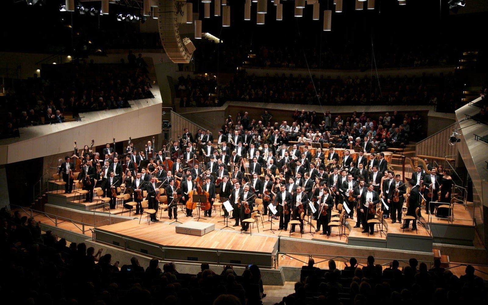 Orchestra members stand for applause on a concert hall stage surrounded by a full audience