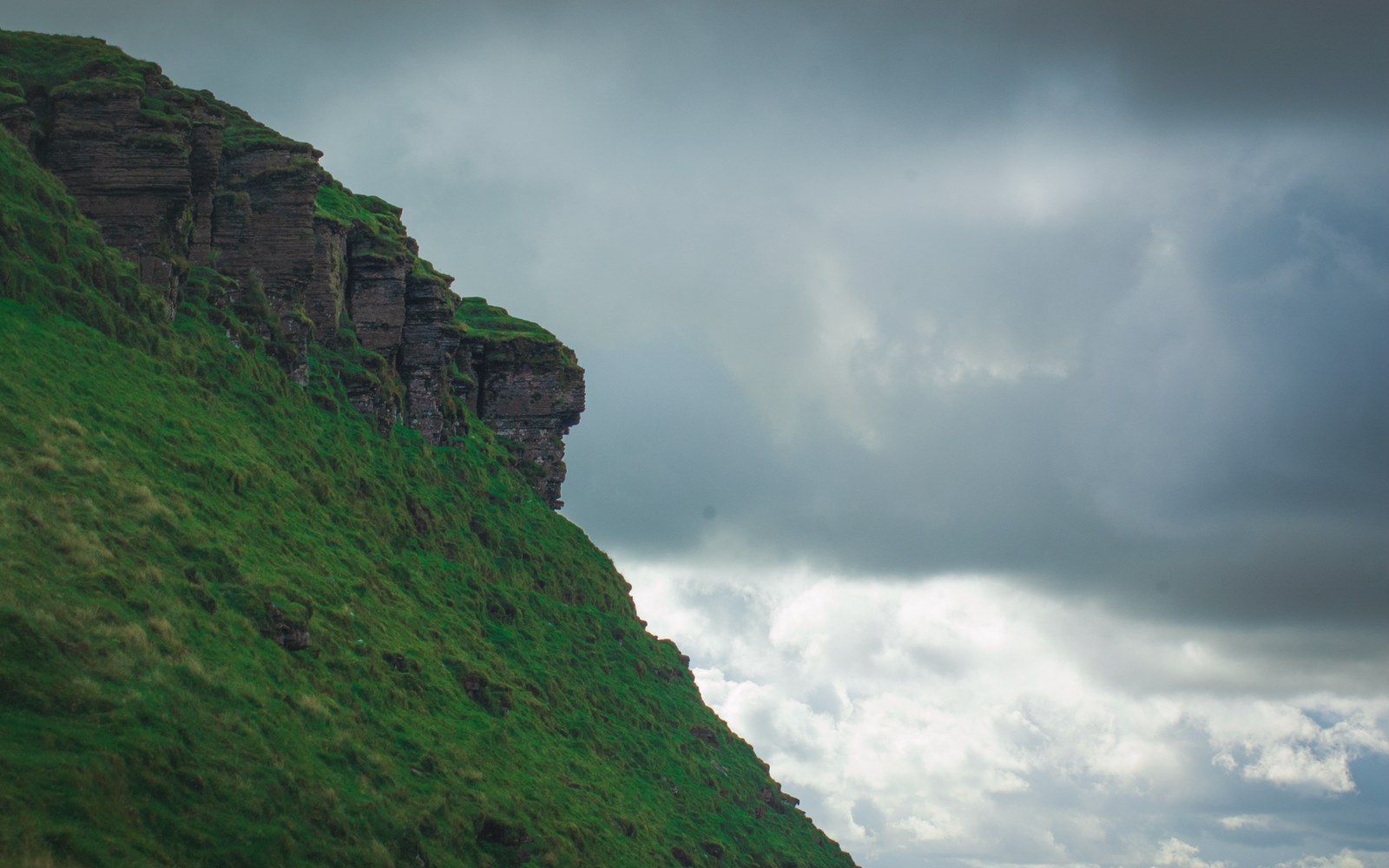 Grass-covered cliff edge with layered rock strata under a heavy, cloudy sky