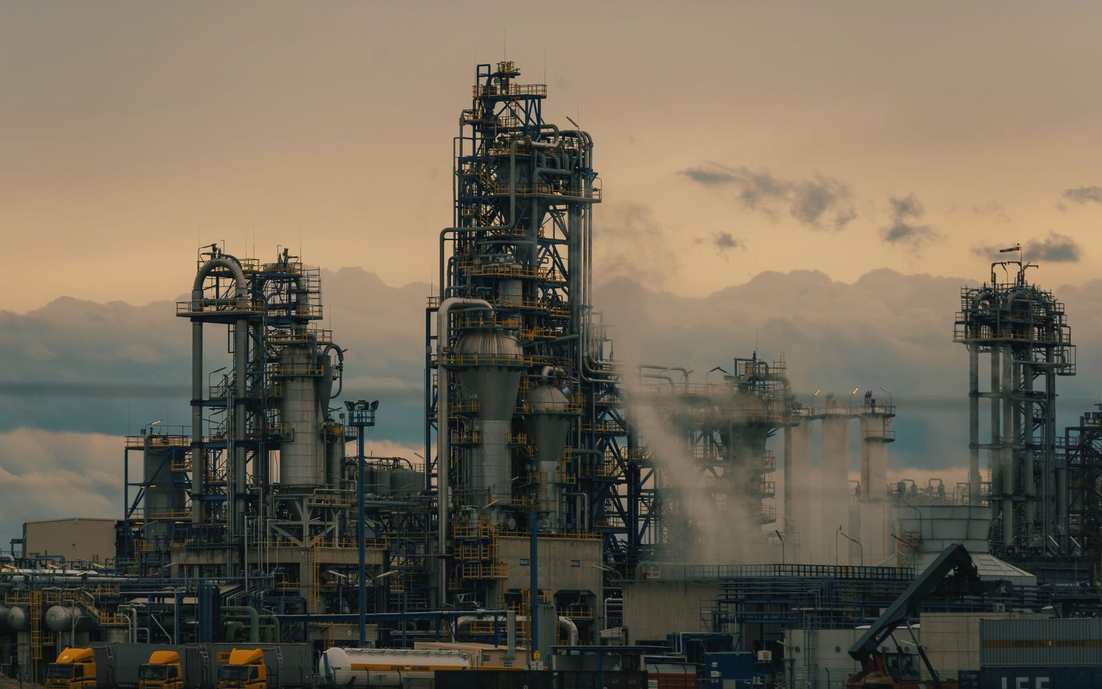 Steam rises from towers and pipework at an oil refinery under a hazy sunset sky