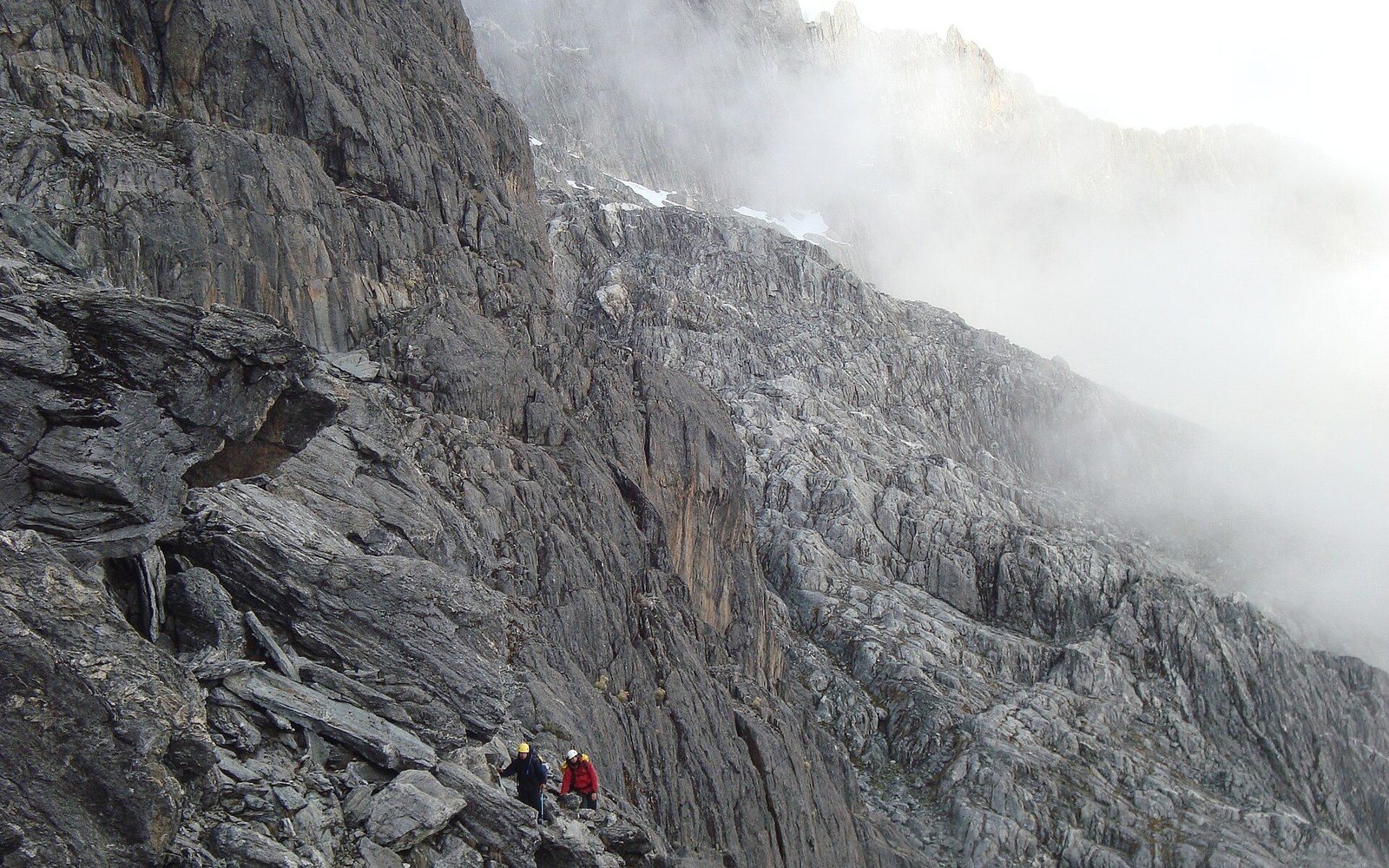 Climbers wearing helmets ascend a steep rocky ridge as clouds drift over the summit