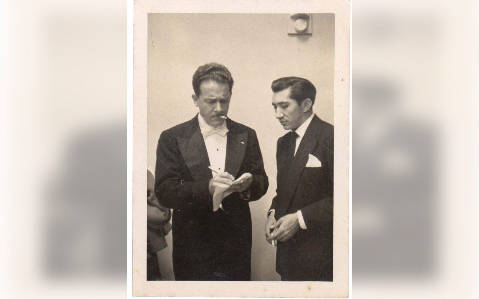Two men in formal evening wear, one writing on a notepad while the other looks on
