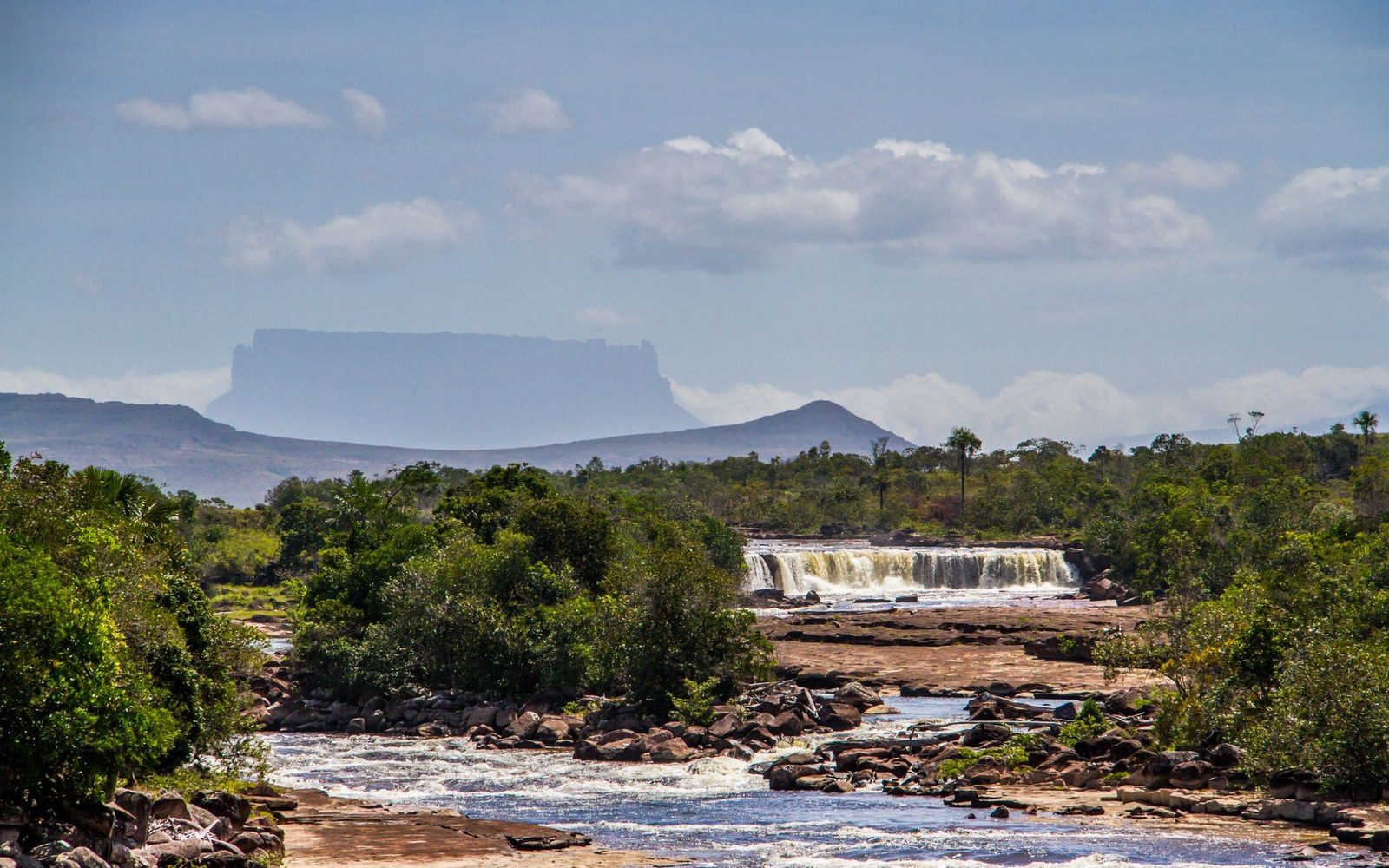 A tabletop tepui rises above savanna forest and a rocky river waterfall
