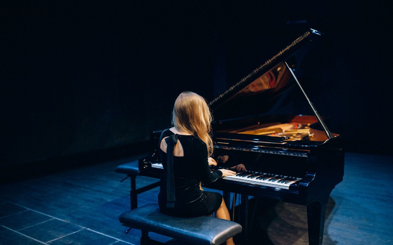A woman in a black dress plays a grand piano alone on a dark stage